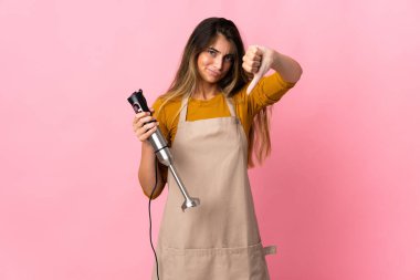 Genç aşçı kadın pembe arka planda izole edilmiş el blenderi kullanıyor.