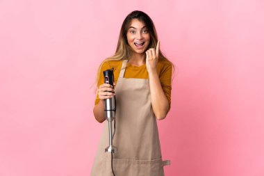 Genç aşçı kadın pembe arka planda izole edilmiş el blenderi kullanıyor. Çözümü bulmak için parmağını kaldırıyor.