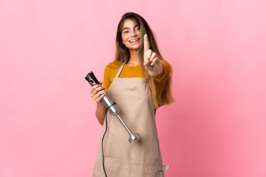 Genç aşçı kadın pembe arka planda izole edilmiş el blenderi kullanıyor ve parmağını kaldırıyor.