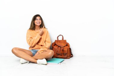 Yerde dizüstü bilgisayarlı genç bir bayan oturuyor. Beyaz arka planda izole edilmiş. Harika bir fikre işaret ediyor.