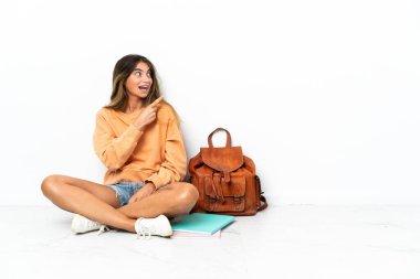 Genç bir kız öğrenci, dizüstü bilgisayarını beyaz arka planda izole bir şekilde yerde oturuyor. Çözümü bulmak için parmağını kaldırıyor.