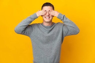 Sarı arka planda izole edilmiş genç beyaz bir adam elleriyle gözlerini kapatıyor ve gülümsüyor.
