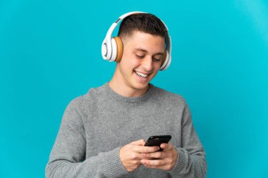 Kafkasyalı genç adam mavi arka planda izole edilmiş müzik dinliyor ve cep telefonu arıyor.
