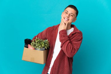 Genç beyaz adam, mavi arka planda izole edilmiş şeylerle dolu bir kutuyu alırken mutlu ve gülümsüyor.