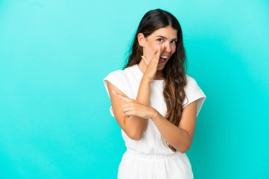 Mavi arka planda izole edilmiş genç beyaz kadın bir ürünü sunmak ve bir şeyler fısıldamak için kenarı işaret ediyor.