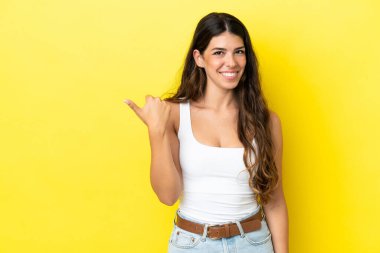 Sarı arka planda izole edilmiş genç beyaz kadın bir ürünü sunmak için kenarı işaret ediyor.