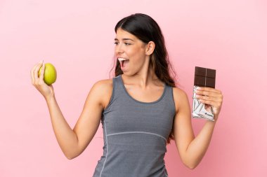 Pembe arka planda izole edilmiş beyaz bir kadın bir elinde çikolata tableti, diğerinde elma alıyor.