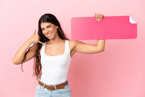 一个被粉色背景隔离的年轻的高加索女人拿着空标语牌 做着电话手势 — 图库照片