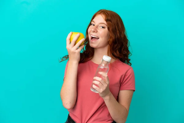 十代の赤毛の女の子はリンゴと水のボトルで青の背景に隔離されました — ストック写真