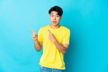 Genç Çinli adam mavi arka planda izole edilmiş korkmuş ve kenarı işaret ediyor.