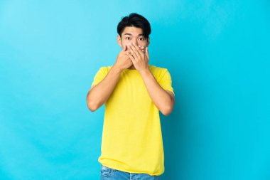 Mavi arka planda izole edilmiş genç Çinli adam elleriyle ağzını kapatıyor.