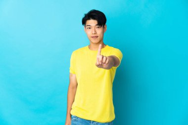 Mavi arka planda izole edilmiş genç bir Çinli parmak kaldırıyor ve gösteriyor.