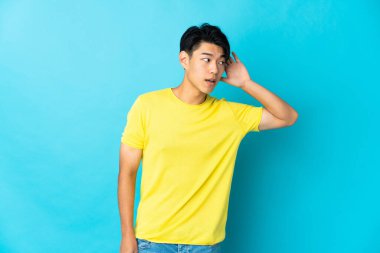 Genç Çinli bir adam mavi arka planda izole edilmiş. Kulağına elini koyarak bir şeyler dinliyor.