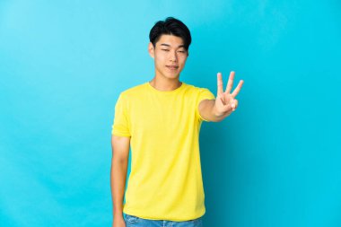 Mavi arka planda izole edilmiş genç bir Çinli mutlu ve parmaklarıyla üçe kadar sayıyor.