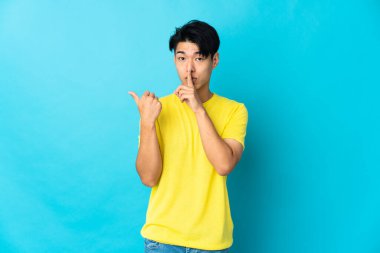 Mavi arka planda izole edilmiş genç bir Çinli yan tarafı işaret ediyor ve sessiz hareketler yapıyor.
