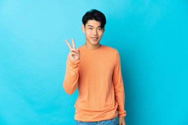 Mavi arka planda izole edilmiş genç Çinli adam gülümsüyor ve zafer işareti gösteriyor.