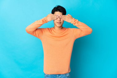 Mavi arka planda izole edilmiş genç bir Çinli. Gözleri elleriyle kaplıyor ve gülümsüyor.