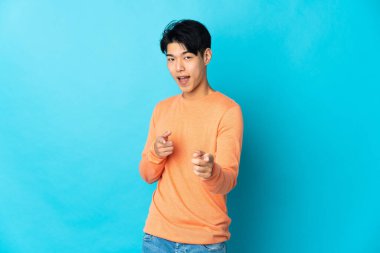 Mavi arka planda izole edilmiş genç bir Çinli ön tarafı işaret ediyor ve gülümsüyor.