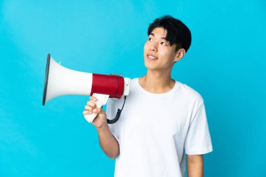 Genç Çinli bir adam mavi arka planda izole edilmiş elinde bir megafonla yukarı bakıyor ve gülümsüyor.