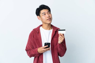 Genç Çinli bir adam beyaz arka planda izole edilmiş. Cep telefonuyla kredi kartıyla alışveriş yapıyor.