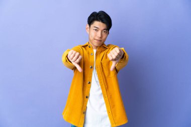 Mor arka planda izole edilmiş genç bir Çinli iki eliyle baş parmağını aşağı gösteriyor.