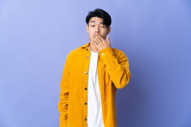 Mor arka planda izole edilmiş genç bir Çinli adam esniyor ve ağzını eliyle açık tutuyor.