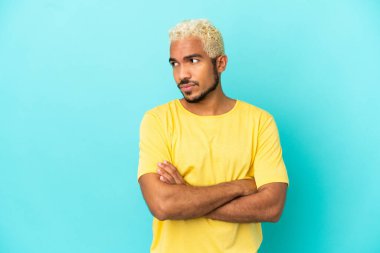 Genç Kolombiyalı yakışıklı adam mavi arka planda izole edilmiş, kollarını kavuşturuyor.