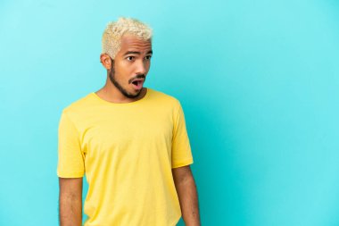 Genç Kolombiyalı yakışıklı adam mavi arka planda izole edilmiş. Yan tarafa bakarken sürpriz jest yapıyor.