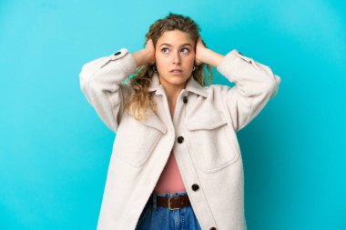 Mavi arka planda izole edilmiş genç sarışın kadın hayal kırıklığına uğramış ve kulaklarını kapatmış.