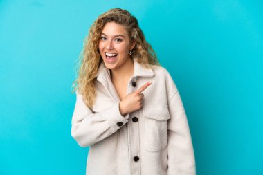 Mavi arka planda izole edilmiş genç sarışın kadın şaşırmış ve işaret ediyor.