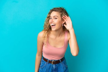 Mavi arka planda izole edilmiş genç sarışın bir kadın. Kulağına elini sürerek bir şeyler dinliyor.