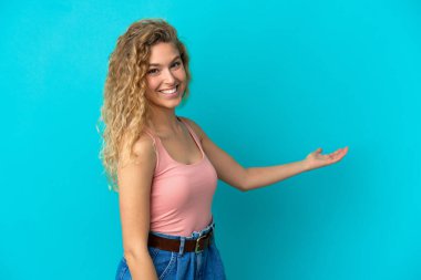 Mavi arka planda izole edilmiş genç sarışın kadın davet etmek için ellerini yana uzatıyor.