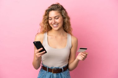 Pembe arka planda izole edilmiş genç sarışın kadın kredi kartıyla cep telefonuyla alışveriş yapıyor.