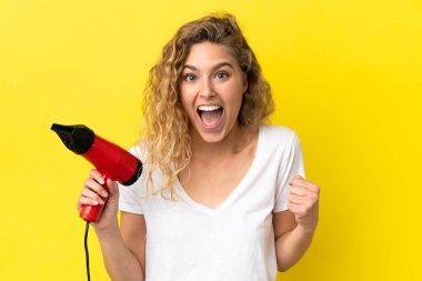 Sarı arka planda izole edilmiş saç kurutma makinesini tutan genç sarışın kadın zaferi kutluyor.