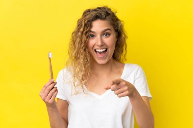 Sarı arka planda dişlerini fırçalayan genç beyaz kadın şaşırmış ve ön tarafı işaret ediyor.