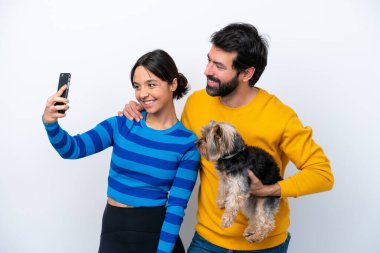 Beyaz arka planda izole edilmiş bir köpeği tutan genç İspanyol kadın cep telefonuyla selfie çekiyor.