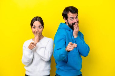 Sarı arka planda izole edilmiş genç beyaz bir çift birini işaret ediyor ve gülüyor.