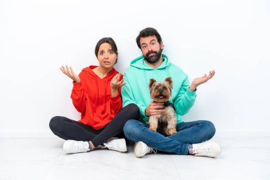 Beyaz arka planda izole edilmiş evcil hayvanlarıyla yerde oturan beyaz bir çift. Şüpheleri var ve kafası karışmış bir ifadeyle.