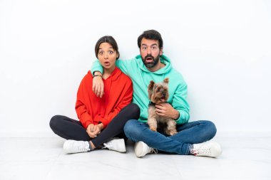 Beyaz arka planda evcil hayvanlarıyla yerde oturan beyaz yüzlü genç bir çift.