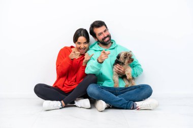 Beyaz arka planda izole edilmiş evcil hayvanlarıyla yerde oturan beyaz tenli genç bir çift kendilerine güvenen bir ifadeyle parmaklarını sana doğrultuyor.
