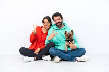 Beyaz arka planda izole edilmiş evcil hayvanlarıyla yerde oturan beyaz bir çift gülümsüyor ve zafer işareti gösteriyor.