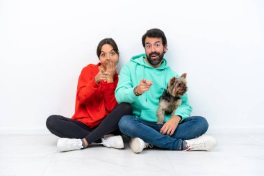 Beyaz arka planda izole edilmiş evcil hayvanlarıyla yerde oturan beyaz bir çift birini işaret ediyor ve gülüyor.