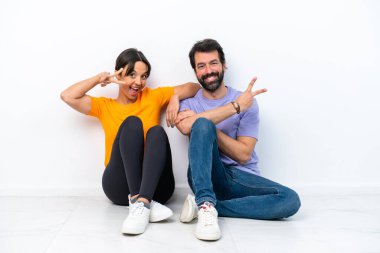Beyaz arka planda izole bir şekilde yerde oturan genç beyaz bir çift gülümsüyor ve zafer işareti gösteriyor.