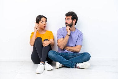 Beyaz arka planda izole bir şekilde yerde oturan genç beyaz bir çift ayakta durup bir fikir düşünüyorlar.