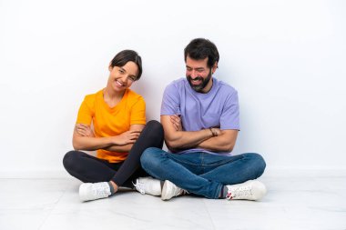 Beyaz arka planda izole bir şekilde yerde oturan beyaz tenli genç çift gülümseyerek kollarını kavuşturuyor.