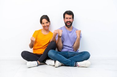 Beyaz arka planda izole bir şekilde yerde oturan beyaz tenli genç çift galibiyetlerini kutluyorlar.