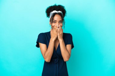 Mavi arka planda izole edilmiş genç Latin kadın mutlu ve gülümsüyor elleriyle ağzını kapatıyor.