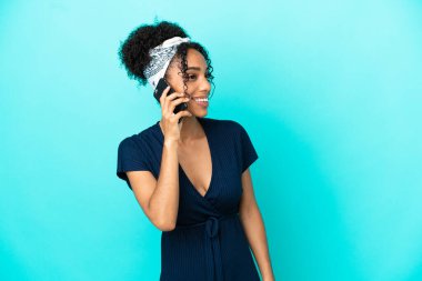 Mavi arka planda izole edilmiş genç Latin bir kadın cep telefonuyla sohbet ediyor.