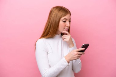 Genç kızıl saçlı kadın pembe arka planda izole edilmiş düşünüyor ve bir mesaj gönderiyor.