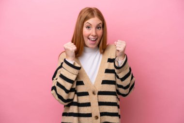 Pembe arka planda izole edilmiş genç kızıl saçlı kadın galibiyeti kutluyor.